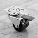 75mm twin wheel braked top plate castor with black polypropylene wheels and grey foot brake on light grey marble, 45 degree three-quarter view from a steep high angle