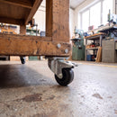80mm braked castor with black rubber wheel bolted under a wooden mobile workbench in a workshop