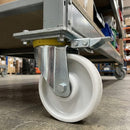 Directional lock swivel castor with white nylon wheel fitted under a grey warehouse shelf trolley